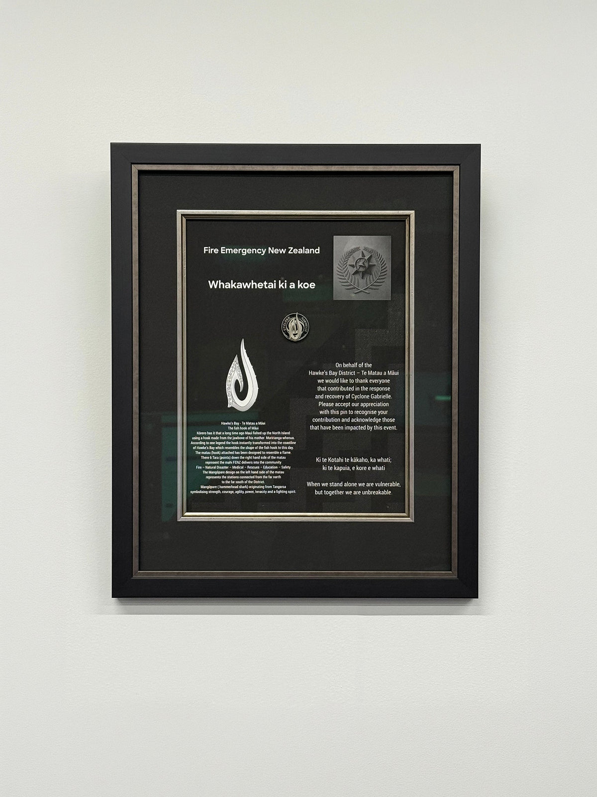 A framed emergency response volunteer pin awarded for cyclone Gabrielle recovery efforts. In a custom made frame mounted on a light gray wall..