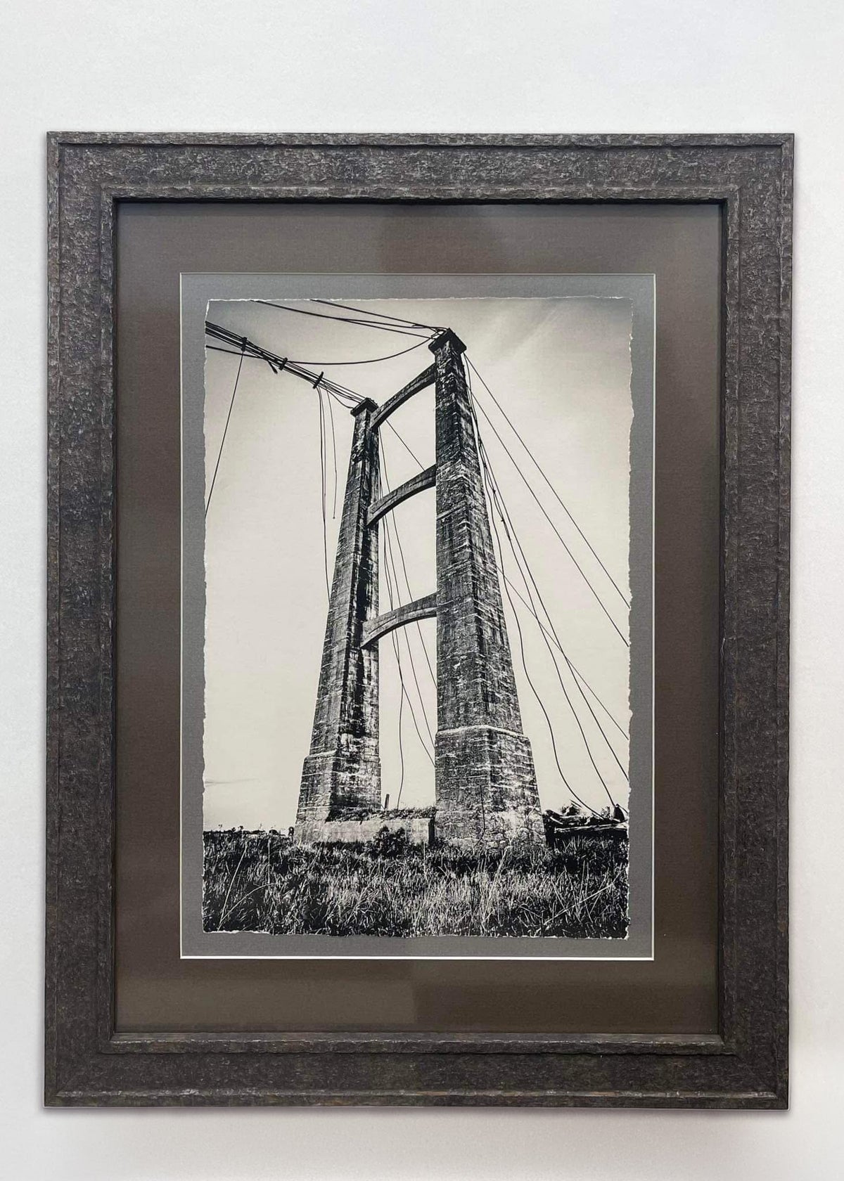 A framed black and white photographic print of the opiki toll  bridge, featuring a dark textured frame and a dark brown mat board, mounted on a plain white wall.
